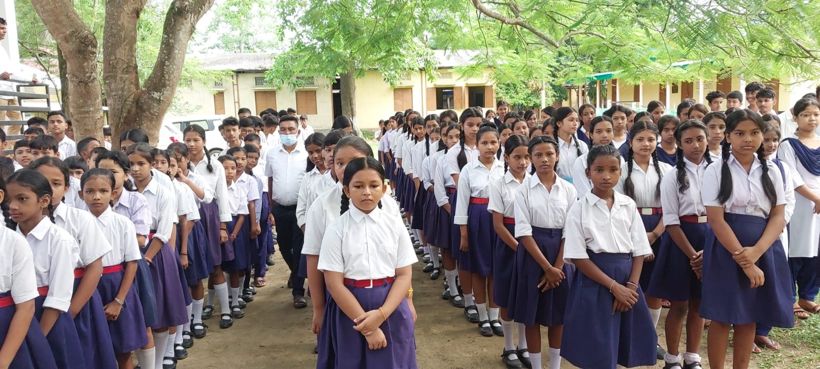 Rows of students in white shirts and navy skirts and trousers at morning assembly under the trees