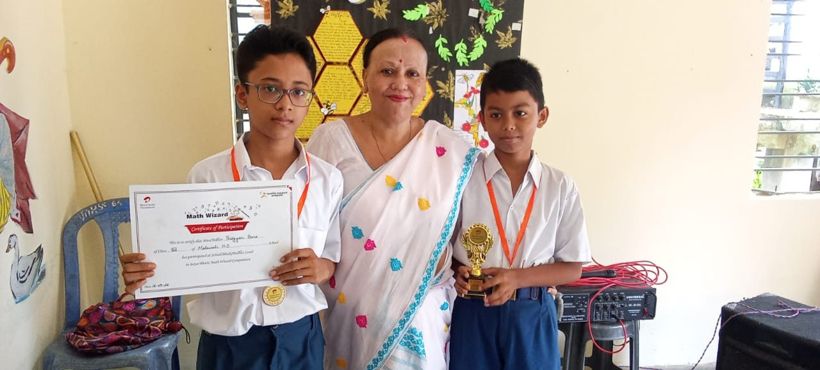 Two students holding a Math Wizard certificate and a trophy, with Principal Shyamalima Borah between them
