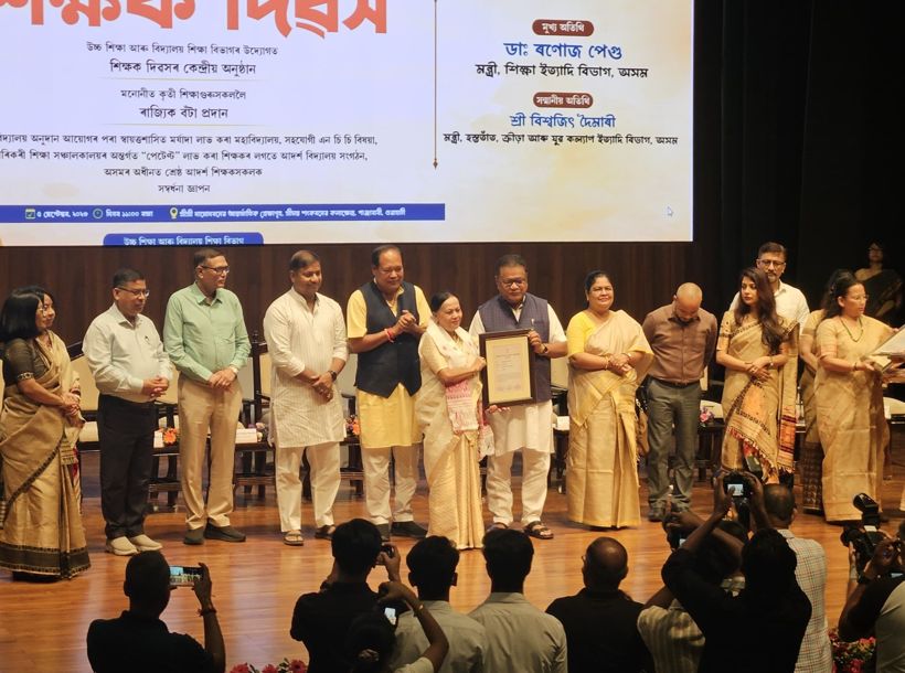 Shyamalima Borah holding her State Teachers’ Award certificate on stage, among other awardees and guests, under a Teachers’ Day banner